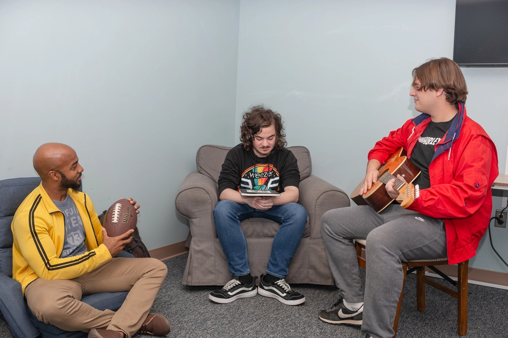 Three friends talking with football and guitar indoors.
